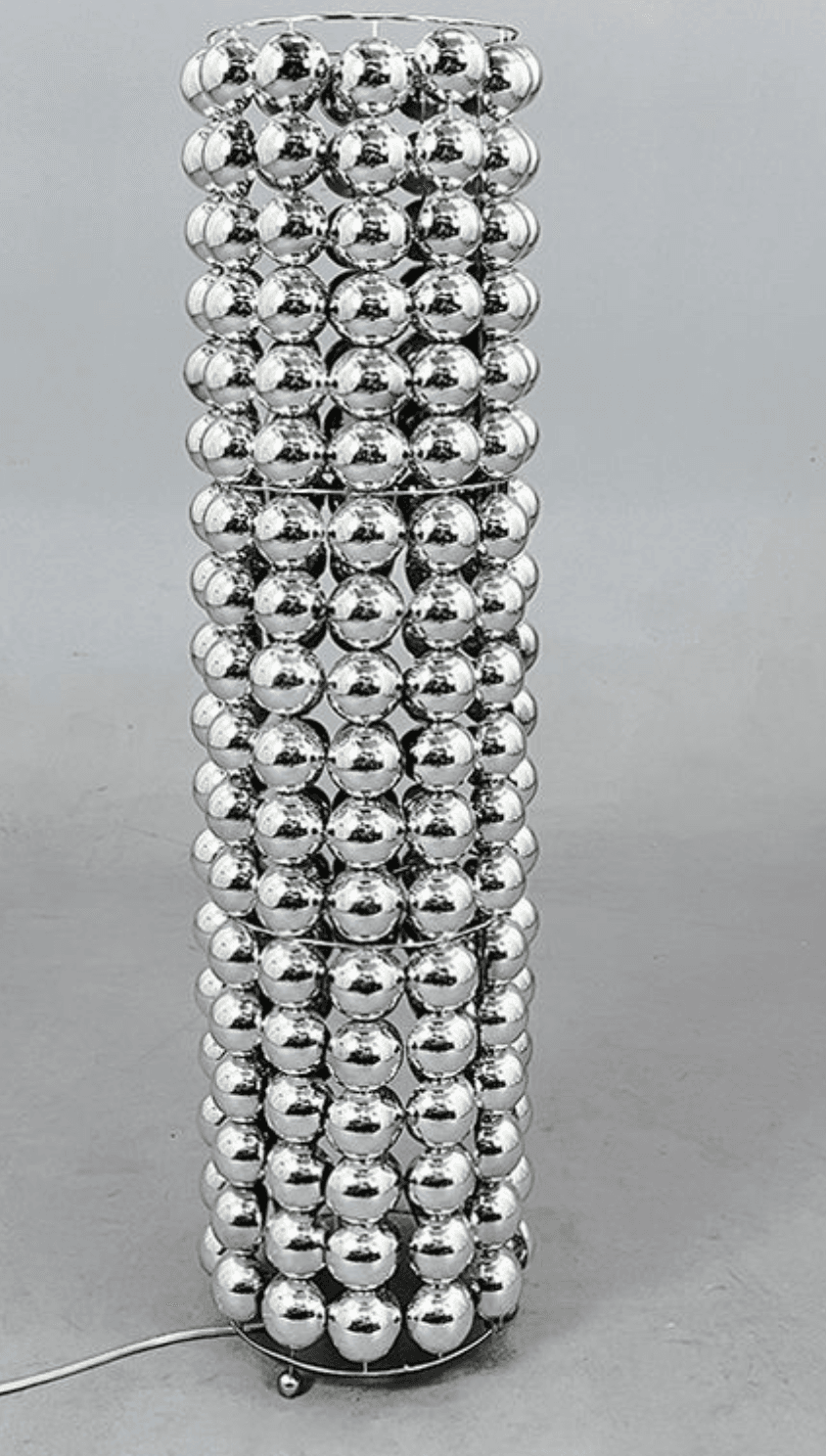 A tall, cylindrical floor lamp with a unique shade constructed from numerous small, spherical, reflective elements. The shade appears to be made of many rows of interconnected silver spheres, creating a textured and decorative surface. The base is minimal, with small feet, and a power cord is visible. — 3D model