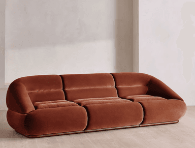 A contemporary three-seater sofa characterized by its rounded, curvilinear form and plush terracotta-colored velvet upholstery. The seating area and backrest are segmented, creating distinct, soft cushions that contribute to its inviting appearance. The sofa features a low profile with a hidden base, giving it a floating aesthetic. Its design suggests a blend of modern comfort with a hint of 1970s lounge aesthetics. — 3D model