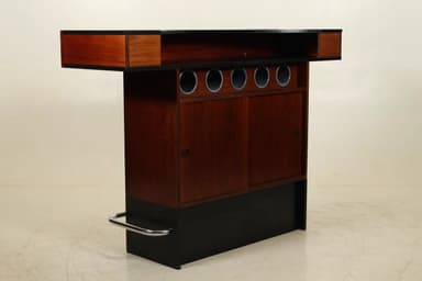 A freestanding bar unit featuring a rectangular top supported by a rectangular pedestal base. The top appears to be made of a dark reddish-brown wood veneer or laminate. The base is primarily black with a metallic (likely chrome) footrest.  The overall design is minimalist and modern in style. The construction suggests a possibly composite or engineered wood material. — 3D model