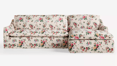 A cream-colored sofa upholstered in a floral fabric with a pattern of roses, peonies, and other flowers in shades of pink, yellow, green, and red. The sofa features a low profile with a slightly sloped armrest. The upholstery covers the entire structure, including the base, cushions, and back. — 3D model