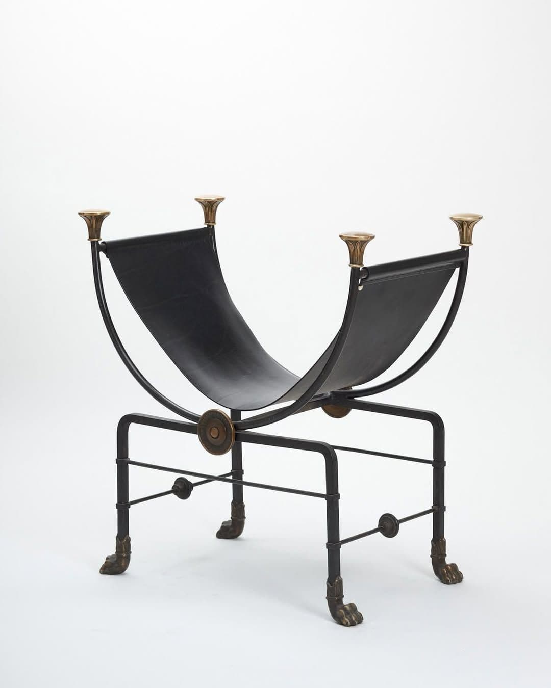 A decorative log holder or magazine rack featuring a black wrought iron or steel frame with brass accents. It has a deep, U-shaped black leather sling for holding items, supported by four legs with ornate brass lion's paw feet and decorative brass finials at the top. — 3D model