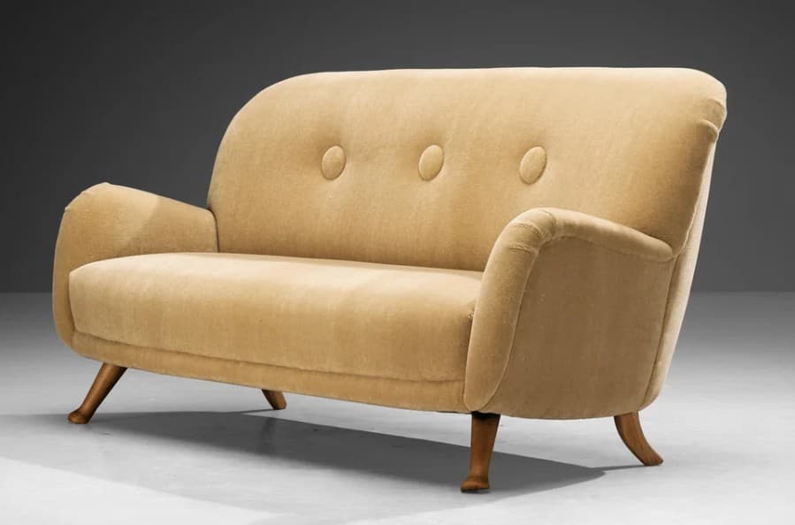 A two-seater sofa with a high, curved backrest and rounded arms. The upholstery appears to be a textured fabric, possibly velvet or chenille, in a light beige or tan color. The backrest features three prominent buttons creating a tufted effect. The sofa is supported by four angled wooden legs. — 3D model