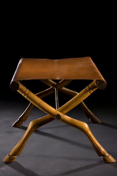 A campaign-style folding stool featuring an X-shaped wooden frame with carved details and a tan leather sling seat. The legs are notably carved to resemble animalistic or hoofed forms, adding an exotic touch. The frame is joined by visible metal hardware at the pivot points and where the leather seat attaches. — 3D model