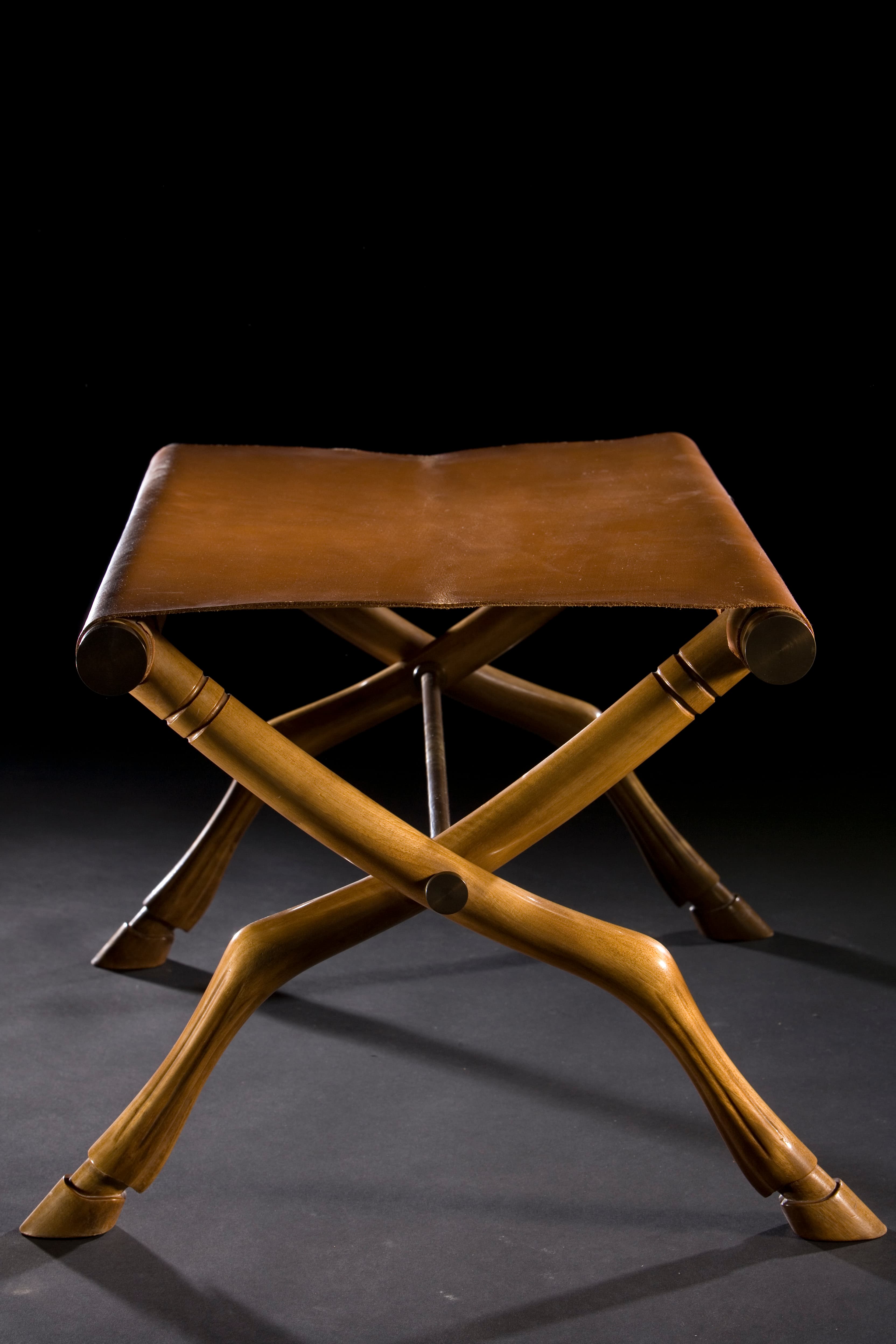 A campaign-style folding stool featuring an X-shaped wooden frame with carved details and a tan leather sling seat. The legs are notably carved to resemble animalistic or hoofed forms, adding an exotic touch. The frame is joined by visible metal hardware at the pivot points and where the leather seat attaches. — 3D model