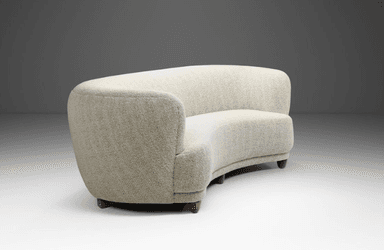 A curved, fully upholstered sofa featuring a serpentine form, rounded back and integrated armrests, and short, tapered wooden legs. It is covered in a light-colored, textured woven fabric. — 3D model