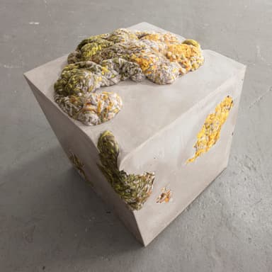 A cubic concrete ottoman with an irregular, textured top surface composed of braided and knotted yarn in shades of yellow, green, and grey. The concrete body features several irregular voids and openings, some of which reveal the same yarn material embedded within. — 3D model