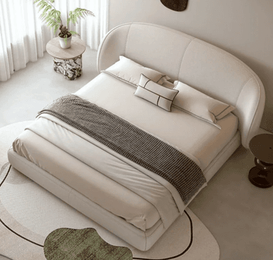 A contemporary upholstered bed with a curved headboard. The bed frame is low-profile and appears to be made of a light-colored fabric. It is dressed with white linens, a grey throw blanket, and decorative pillows. A small, organically shaped side table with a marble-patterned top and a sculptural base is placed to the left of the bed. To the right, a small, dark-colored stool-like side table is visible. A rug with abstract green and beige shapes is partially in view in the foreground. — 3D model