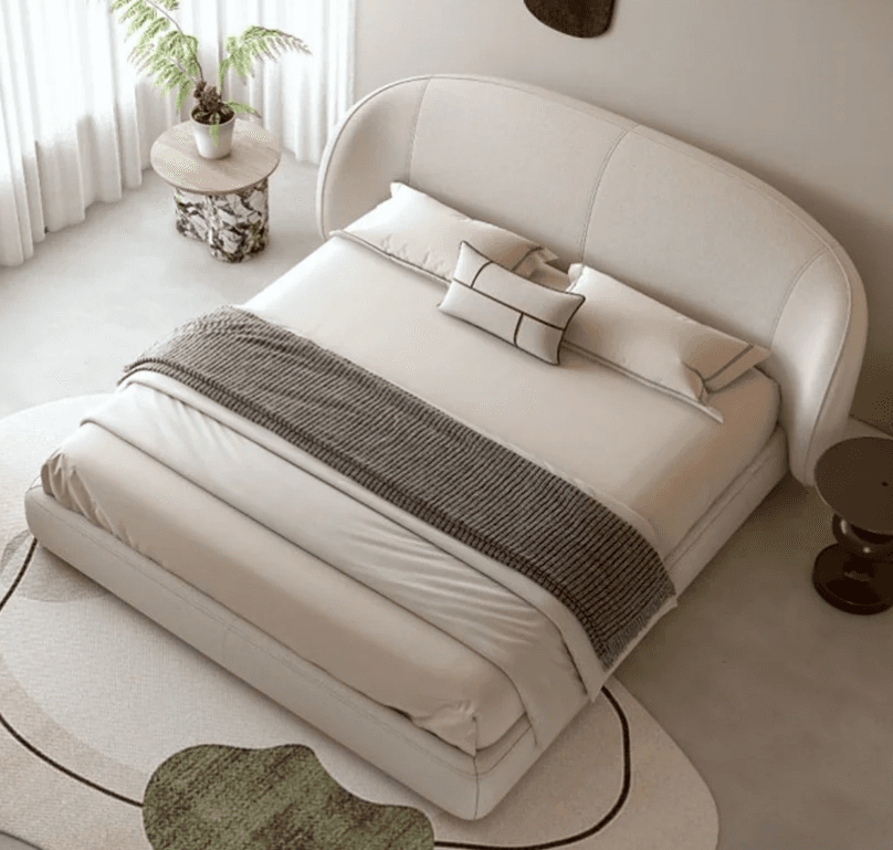 A contemporary upholstered bed with a curved headboard. The bed frame is low-profile and appears to be made of a light-colored fabric. It is dressed with white linens, a grey throw blanket, and decorative pillows. A small, organically shaped side table with a marble-patterned top and a sculptural base is placed to the left of the bed. To the right, a small, dark-colored stool-like side table is visible. A rug with abstract green and beige shapes is partially in view in the foreground. — 3D model
