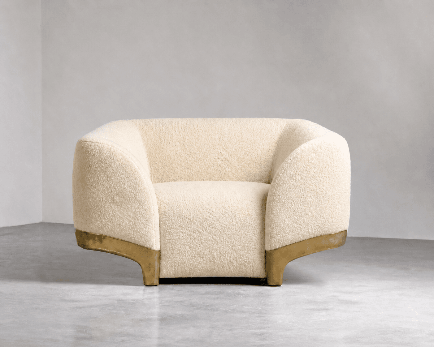 A cream-colored, rounded armchair upholstered in a textured boucle fabric. It features a low profile with wide, curved armrests that blend into the seat and back. The base of the armchair is accented with a curved brass or gold-toned metal trim that extends down to form the legs. — 3D model