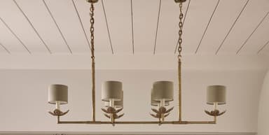 A linear chandelier suspended by two chains. The fixture has a horizontal bar with six lights, each featuring a white cylindrical lampshade supported by a decorative metal arm with leaf-like accents. The metal frame has a distressed gold finish. — 3D model