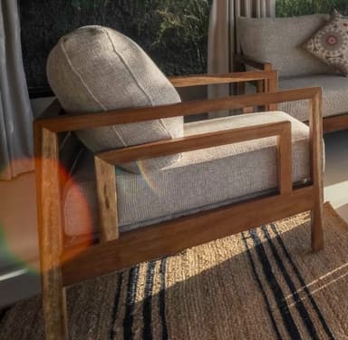 A contemporary armchair featuring a robust, rectilinear wooden frame in a natural finish, complemented by plush, light-beige fabric cushions for the seat and backrest. The design is clean and minimalist, highlighting the natural grain of the wood. — 3D model