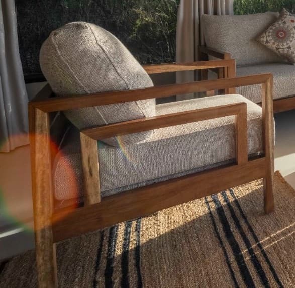 A contemporary armchair featuring a robust, rectilinear wooden frame in a natural finish, complemented by plush, light-beige fabric cushions for the seat and backrest. The design is clean and minimalist, highlighting the natural grain of the wood. — 3D model