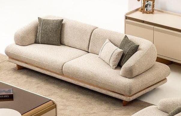 A contemporary low-profile sofa with a distinctive curved design, upholstered in a light beige textured fabric. It features a continuous, gently curved backrest and armrests, three seat cushions, and is supported by a recessed wooden base with visible trim along the bottom edge of the upholstery. Two decorative throw pillows in a darker gray fabric with light piping are included. — 3D model