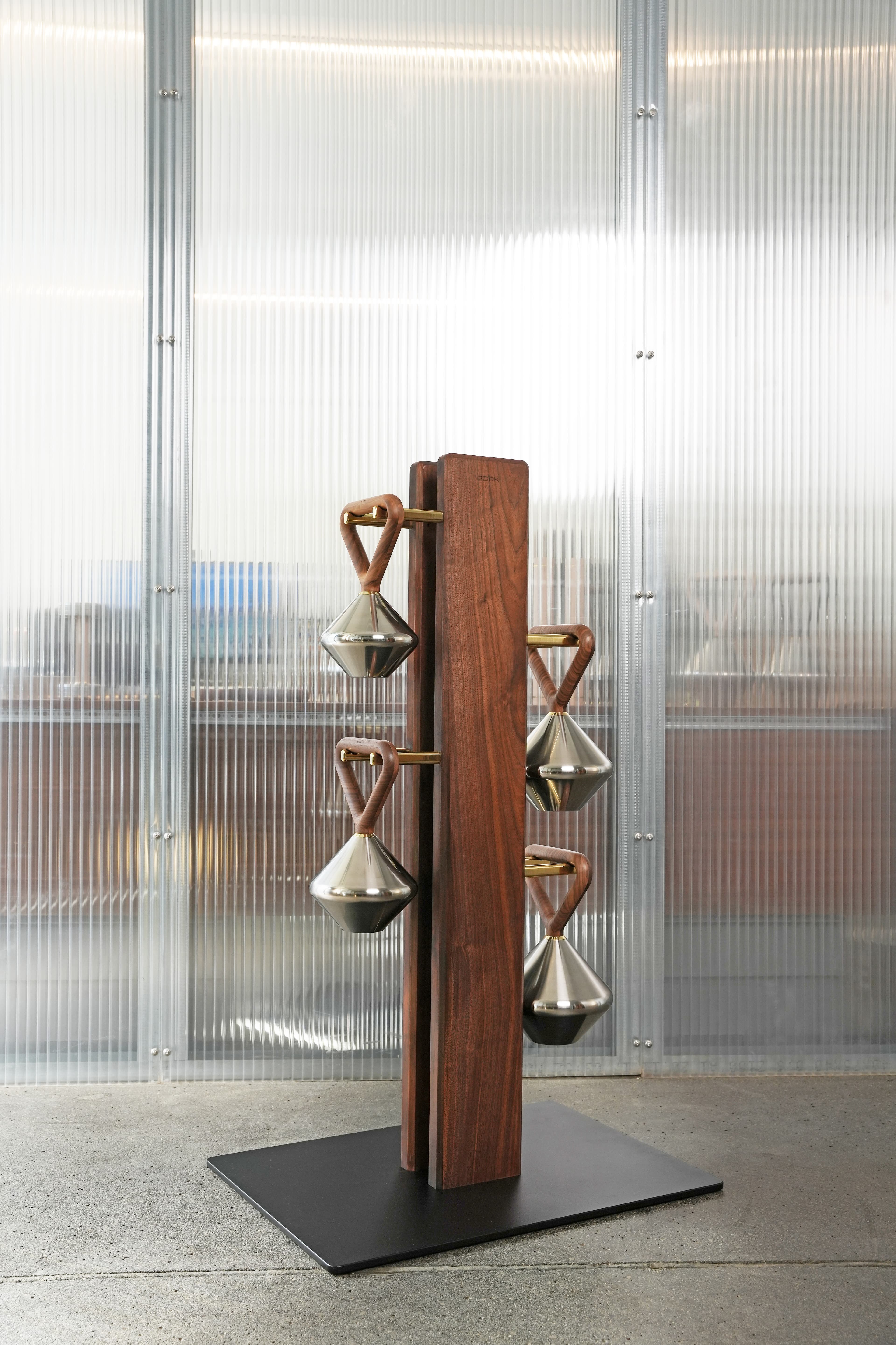 A modern freestanding coat rack featuring two vertical walnut wood columns, from which six conical polished metal hangers are suspended by brass rods and curved wooden grips. It stands on a flat, black square metal base. — 3D model