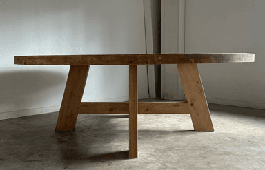 A round dining table with a thick, solid wood top and a substantial base constructed from heavy timber. The base features four angled legs connected by a cross-brace structure, providing stability and a rustic aesthetic. — 3D model