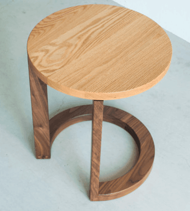 A modern end table with a circular light wood top and a U-shaped base made of dark wood. The base is constructed from two vertical supports connected by a curved element that forms the lower part of the stand. The materials appear to be solid wood with a natural finish. — 3D model