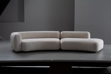 A two-piece modular sofa featuring a distinct curved silhouette and upholstered in a light-colored, textured boucle fabric. The design is contemporary with rounded edges and a low profile, comprising a left arm section and a right armless chaise-like section that together form a gentle curve. The sofa sits close to the floor on concealed or minimalist feet. — 3D model