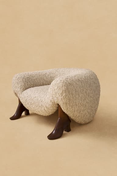 An eccentric upholstered armchair featuring a rounded, organic form and four dark wooden legs sculpted to resemble high-heeled boots. The chair is covered in a textured, light beige boucle fabric, providing a soft and inviting appearance. The design is whimsical and sculptural, making it a distinctive statement piece. — 3D model