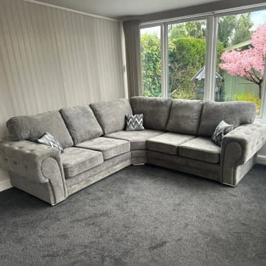 A large, L-shaped sectional sofa upholstered in light gray fabric, featuring button tufting on the rolled arms and base, loose seat and back cushions, and supported by low chrome block feet. Two patterned throw pillows are included. — 3D model