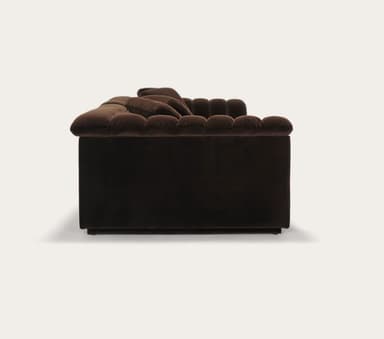 A long, three-seater sofa with a channeled design on the backrests and armrests. The sofa appears to be upholstered in a plush fabric and features a low profile with a distinct armrest that curves outwards. — 3D model