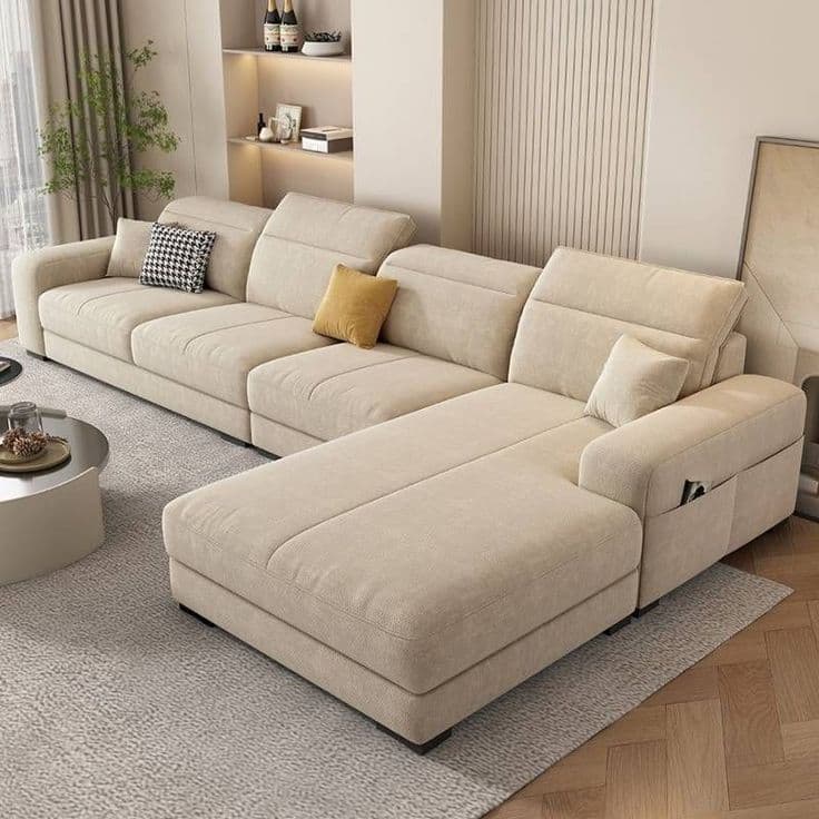 A modern, modular L-shaped sectional sofa upholstered in light beige fabric, featuring multiple seating sections including a prominent chaise lounge. The backrests are equipped with adjustable headrests for enhanced comfort, and the armrest of the chaise section includes integrated storage pockets. The sofa rests on low-profile dark feet, presenting a clean and contemporary aesthetic. — 3D model