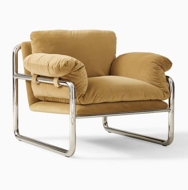 A modern armchair featuring a polished chrome tubular frame with large, plush, camel-colored fabric cushions. The frame forms a continuous loop for the base and back, with horizontal bars supporting the seat and providing structure for the armrests. The seat, back, and armrest cushions are soft and oversized, creating a comfortable, inviting appearance. The armrest cushions are secured to the frame with fabric straps. — 3D model