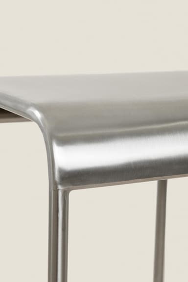 A close-up image displaying the upper portion of a backless stool crafted from brushed metal. The seat features a smooth, gently curved design that transitions seamlessly into the supporting structure, likely cylindrical legs, partially visible. The material exhibits a refined, matte silver finish with a distinct brushed texture. — 3D model