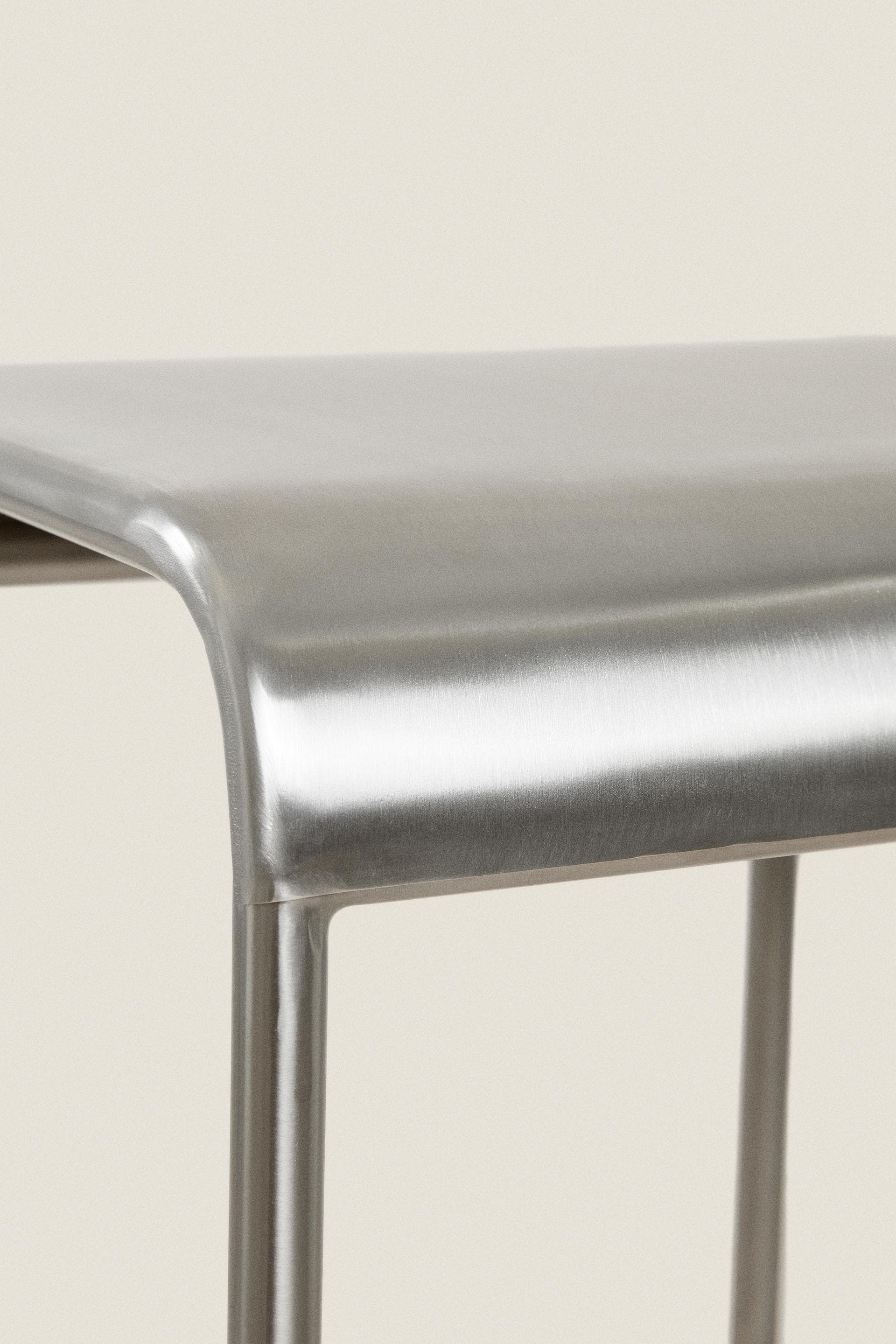A close-up image displaying the upper portion of a backless stool crafted from brushed metal. The seat features a smooth, gently curved design that transitions seamlessly into the supporting structure, likely cylindrical legs, partially visible. The material exhibits a refined, matte silver finish with a distinct brushed texture. — 3D model