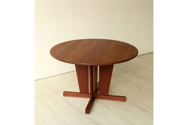 A round wooden table with a sturdy pedestal base, featuring a reddish-brown finish. The top is circular, and the base consists of a central column that flares out into an X-shaped foot structure on the floor, providing stable support. The design appears functional and clean-lined. — 3D model