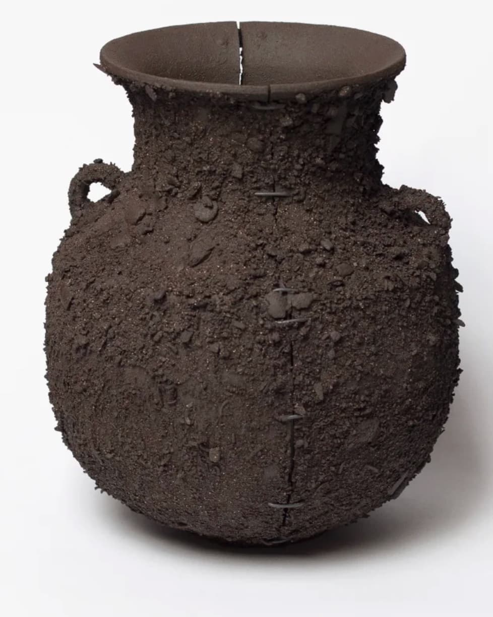 A dark, heavily textured ceramic urn featuring a globular body, a wide, flared rim, and a single visible handle stub. The surface exhibits a coarse, granular texture, reminiscent of rough earth or volcanic rock. A prominent vertical crack is visible along the side, intentionally repaired with multiple metal staples, contributing to a wabi-sabi aesthetic. The overall color is a deep, matte brown or charcoal, and the object appears unglazed. — 3D model