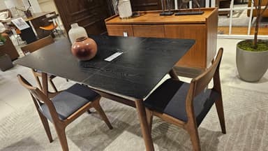 A rectangular dining table featuring a black wood-grain finished top with softly rounded corners. The tabletop appears to be extendable, indicated by visible seams. It stands on four splayed and tapered legs crafted from a contrasting brown wood. — 3D model
