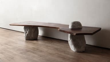 A unique bench featuring a live-edge wooden slab top supported by two irregular, natural stone bases. The wood exhibits a dark brown hue with visible grain and natural imperfections, while the stone bases are light gray and appear rough-hewn, providing a strong textural contrast. The overall design emphasizes natural forms and materials. — 3D model
