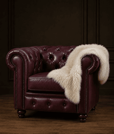 A classic Chesterfield-style armchair, upholstered in rich burgundy leather, featuring deep button tufting on the backrest and rolled arms, and detailed with brass nailhead trim along the front edges and base. The chair sits on dark, turned wooden feet. A cream-colored faux fur throw blanket is draped over one arm and part of the seat. — 3D model