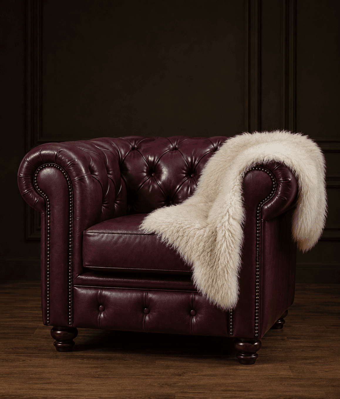 A classic Chesterfield-style armchair, upholstered in rich burgundy leather, featuring deep button tufting on the backrest and rolled arms, and detailed with brass nailhead trim along the front edges and base. The chair sits on dark, turned wooden feet. A cream-colored faux fur throw blanket is draped over one arm and part of the seat. — 3D model