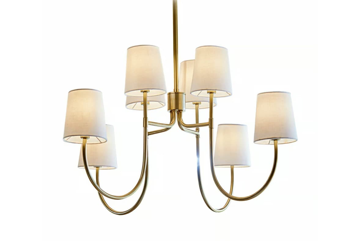 The image displays a contemporary chandelier featuring a brass-finished frame and multiple individual light sources, each covered by a tapered fabric shade. The central stem descends from the top, branching out into gracefully curved arms that support seven lights in total. The shades are an off-white or cream color, providing a soft, diffused light, while the brass structure adds a touch of classic elegance. — 3D model