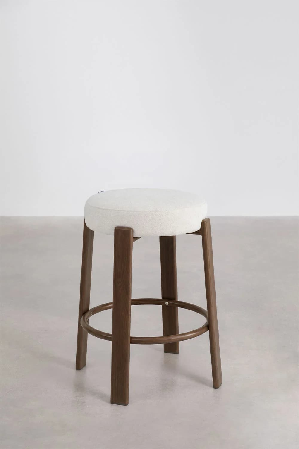 A contemporary bar stool featuring a round, thickly upholstered seat set atop a solid wood base. The four sturdy legs are made of dark brown wood, slightly tapered, and connected by a circular metal footrest for stability and comfort. The seat is covered in an off-white, textured fabric, providing a clean and minimalist aesthetic. — 3D model