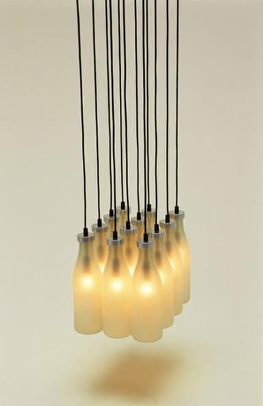 A multi-light cluster pendant chandelier featuring several frosted glass shades, each designed to resemble a classic milk bottle. The bottles are suspended by individual black electrical cords from a ceiling mount (not visible). Each bottle has a silver-tone metallic cap where it connects to the cord, housing the light source. The frosted glass diffuses the light, creating a soft, warm glow. — 3D model