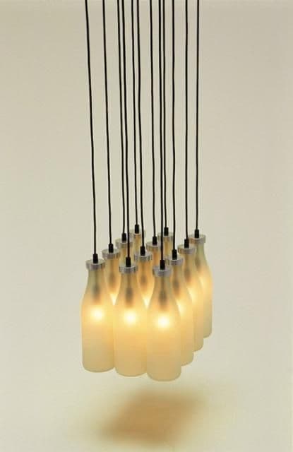 A multi-light cluster pendant chandelier featuring several frosted glass shades, each designed to resemble a classic milk bottle. The bottles are suspended by individual black electrical cords from a ceiling mount (not visible). Each bottle has a silver-tone metallic cap where it connects to the cord, housing the light source. The frosted glass diffuses the light, creating a soft, warm glow. — 3D model