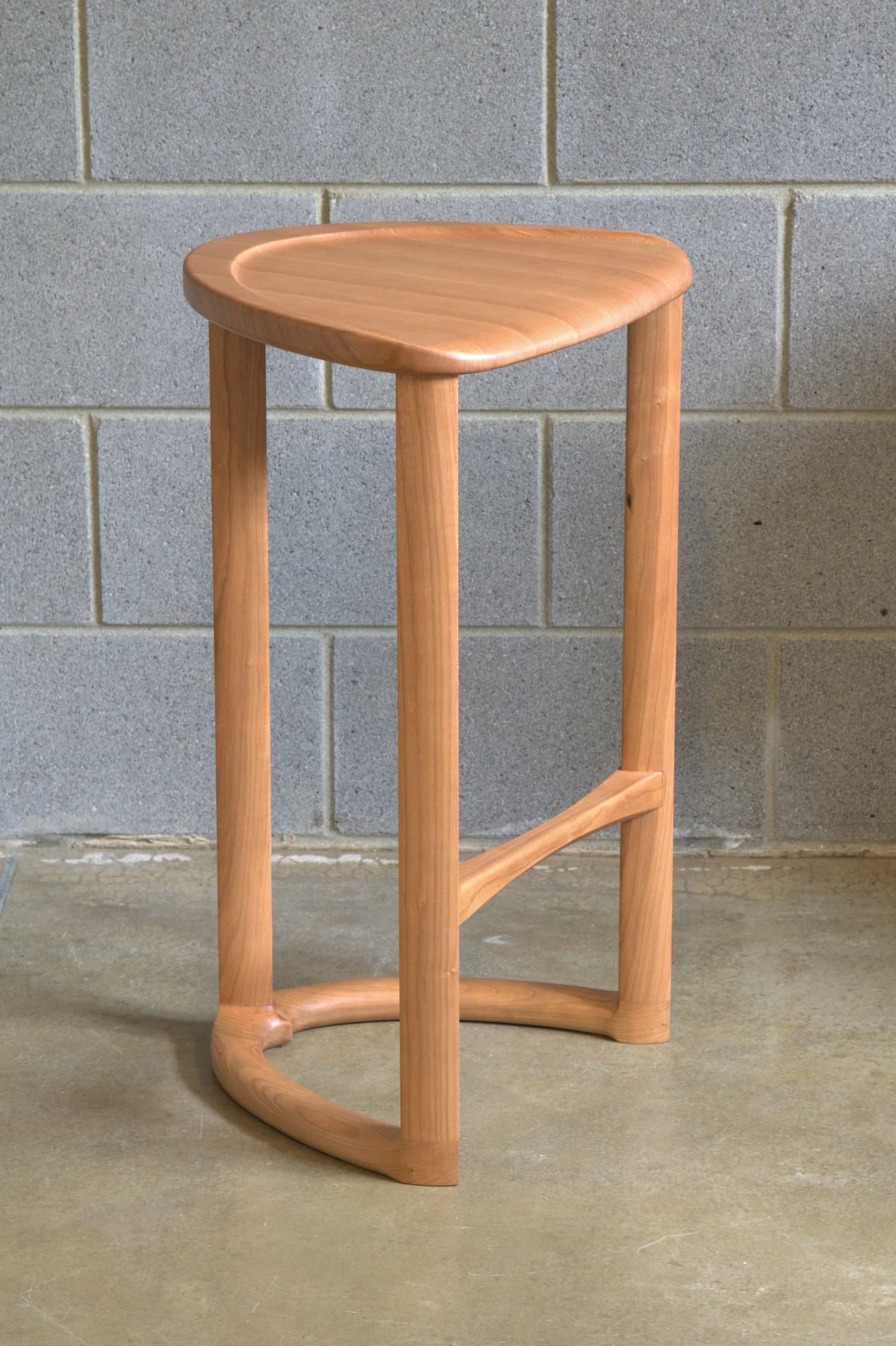 A wooden bar stool with a triangular, slightly dished seat and three cylindrical legs. One leg is positioned at the front and two at the back. A curved stretcher connects the two back legs near the bottom, and a diagonal stretcher connects the front leg to the rear left leg. The stool is constructed from light-colored wood with a visible grain. — 3D model