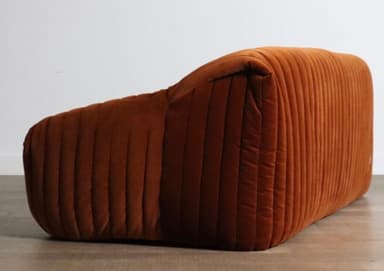 A plush, channel-tufted sofa upholstered entirely in a warm, burnt orange velvet fabric. It features a low-profile design with rounded contours and integrated armrests that seamlessly flow into the backrest and base. The distinctive vertical channel tufting provides a rich texture across all upholstered surfaces. — 3D model