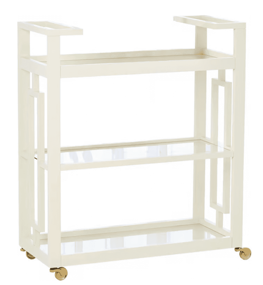 A modern three-tier bar cart featuring a cream-colored metal frame with geometric Art Deco-inspired side panels, clear glass shelves, and brass-finished casters for mobility. — 3D model