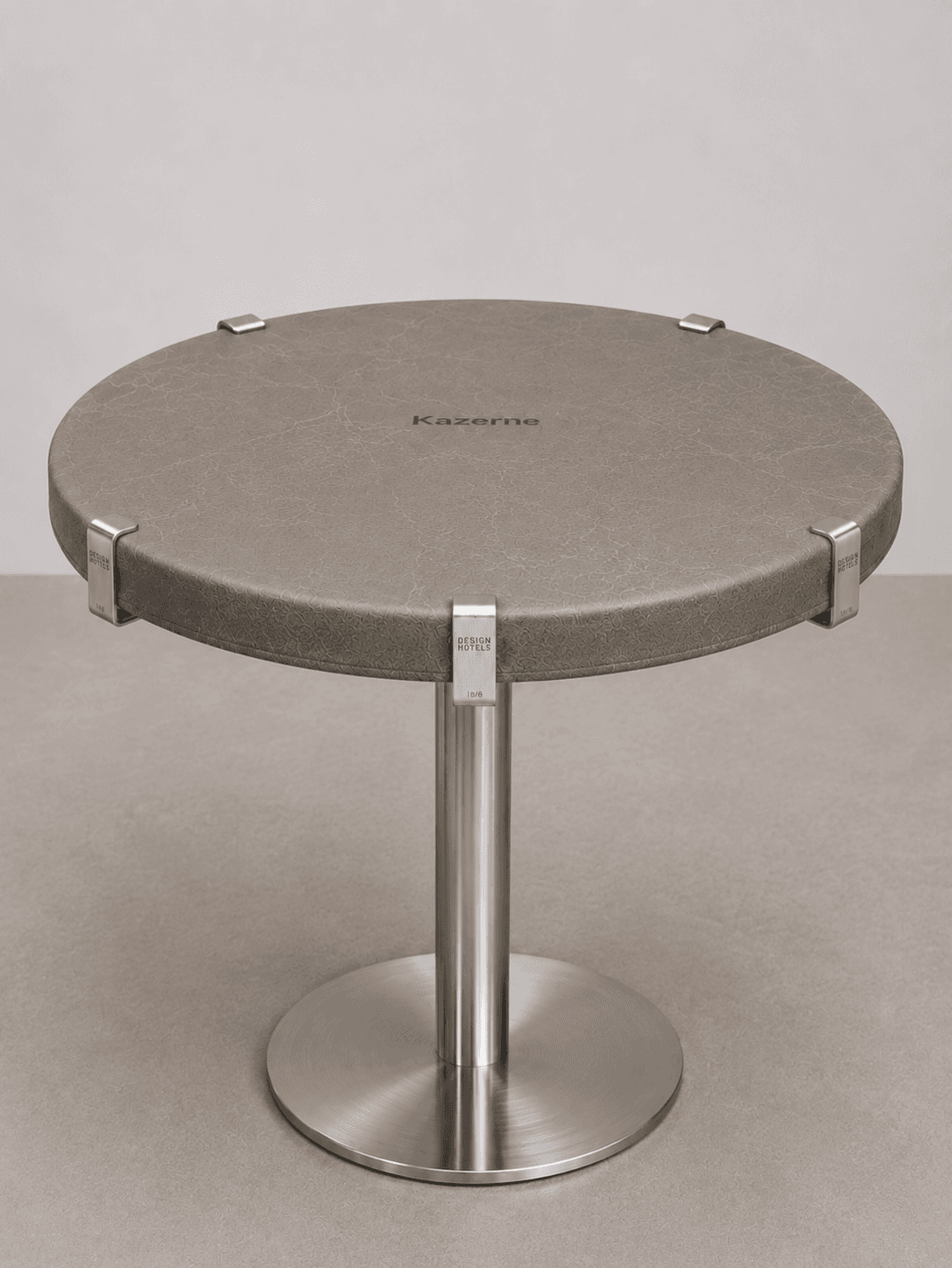 A contemporary round accent table featuring a textured grey upholstered top supported by a central brushed metal pedestal base. The top is secured to the pedestal by four external metal clips evenly spaced around its perimeter. The base consists of a cylindrical column attached to a flat, round metal disc. — 3D model