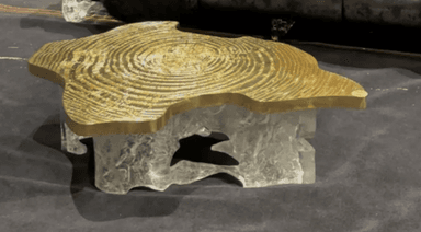 A striking coffee table featuring an irregular, organic-shaped tabletop made of polished golden brass, adorned with a detailed concentric ring pattern. The base is crafted from a chunky, translucent material, likely clear resin or acrylic, sculpted into an abstract, ice-like form, creating a visually compelling contrast with the metallic top. The table exhibits a low profile. — 3D model