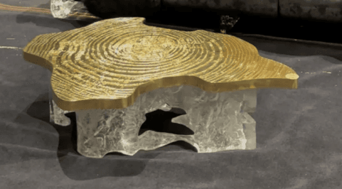 A striking coffee table featuring an irregular, organic-shaped tabletop made of polished golden brass, adorned with a detailed concentric ring pattern. The base is crafted from a chunky, translucent material, likely clear resin or acrylic, sculpted into an abstract, ice-like form, creating a visually compelling contrast with the metallic top. The table exhibits a low profile. — 3D model