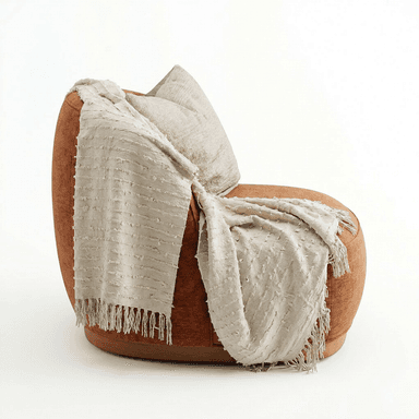 A contemporary armchair with a rounded silhouette, upholstered in a textured, rust-colored fabric. It features a low profile with a visible wooden base. A textured throw blanket with fringes and a decorative pillow are draped over the back and armrest. — 3D model