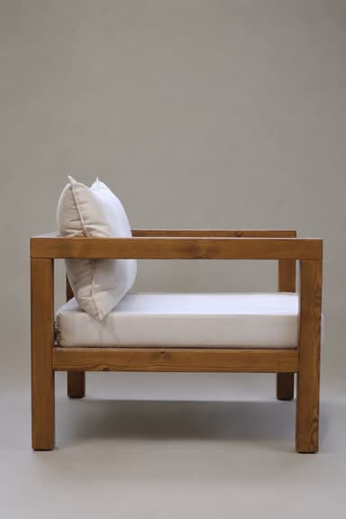 A robust armchair featuring a rectilinear frame constructed from solid, warm-toned wood, likely pine, with visible grain. The chair is equipped with thick, plush, and removable cushions for both the seat and backrest, upholstered in a light, neutral-colored fabric such as beige or off-white. The design emphasizes clean lines and a sturdy construction, presenting a modern rustic or minimalist aesthetic suitable for various indoor or covered outdoor settings. — 3D model