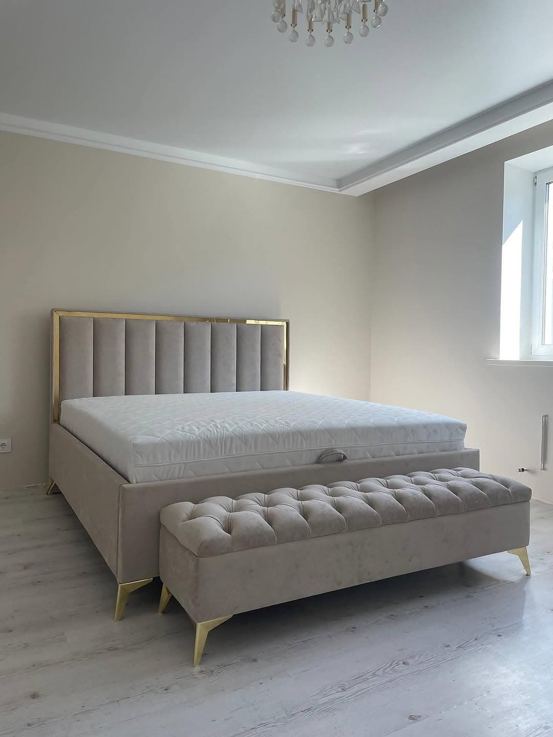 A modern platform bed featuring a tall, rectangular headboard with vertical channel tufting and a gold-tone metal frame border. The bed frame is fully upholstered in a light beige fabric, resting on four slender, angled gold-tone metal legs. A matching upholstered bench with diamond tufting and similar gold-tone legs is positioned at the foot of the bed, completing the set. — 3D model
