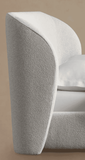 A close-up view of a modern upholstered bed frame, featuring a softly curved, enveloping headboard and side rails covered in a textured, light-colored boucle fabric. A white pillow and bedsheet are visible, indicating its function. — 3D model