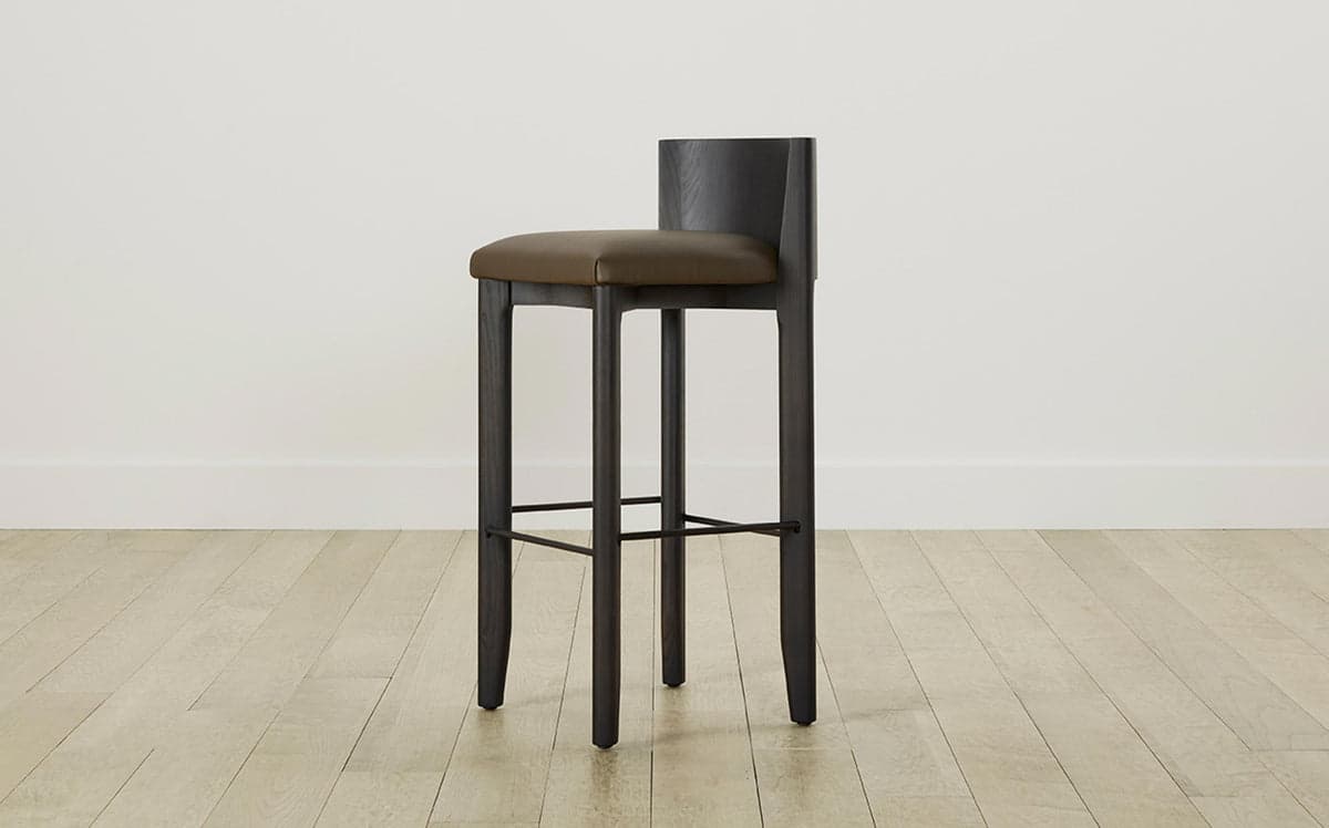 A modern bar stool featuring a dark stained wooden frame and a padded, upholstered seat with a low backrest. The stool has four sturdy, slightly tapered legs and an integrated metal footrest. — 3D model