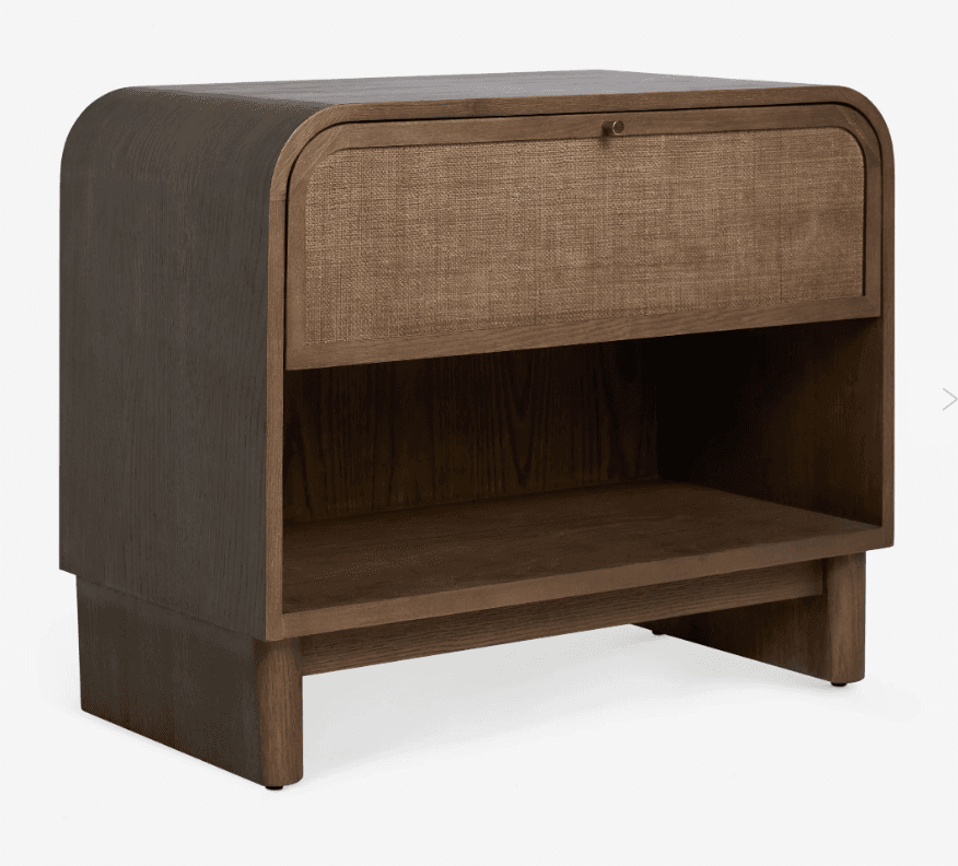 A contemporary nightstand featuring a dark-stained wooden frame with gently rounded top edges. It includes a single drawer with a woven cane front and a small brass knob, positioned above an open lower storage shelf. The base is a solid, plinth-like structure with subtle, short legs. — 3D model